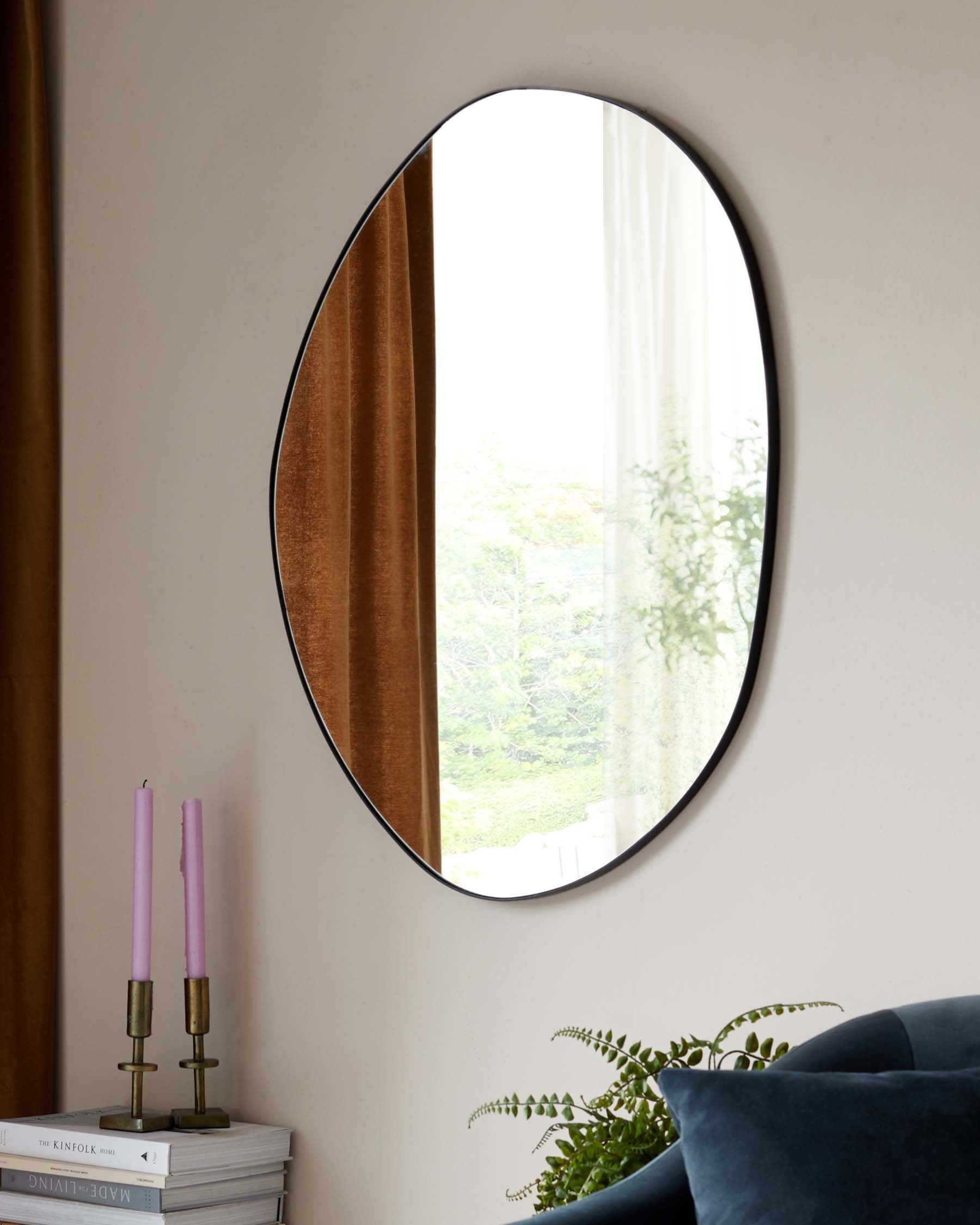 An oval-shaped mirror with a thin black frame mounted on a wall, beside a stack of books topped with a brass candlestick holder with two pink candles. Below, a corner of a blue velvet sofa is visible, with a green potted plant on its arm.