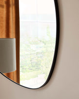 Round wall mirror with a thin black frame, reflecting natural light and greenery from the outside view.