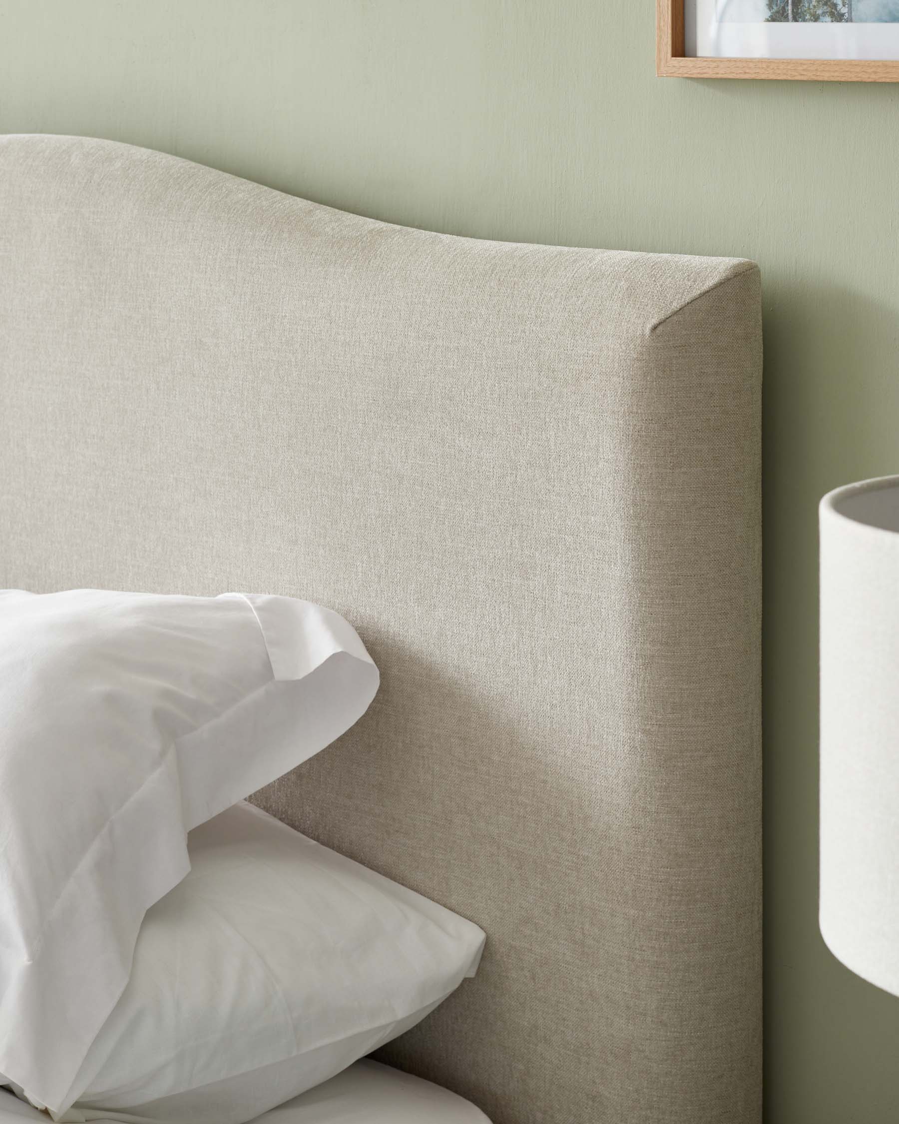 Beige upholstered headboard with a subtle curve at the top edge, complemented by crisp white bedding and set against a soft green wall. A neutral lampshade is partially visible to the side.