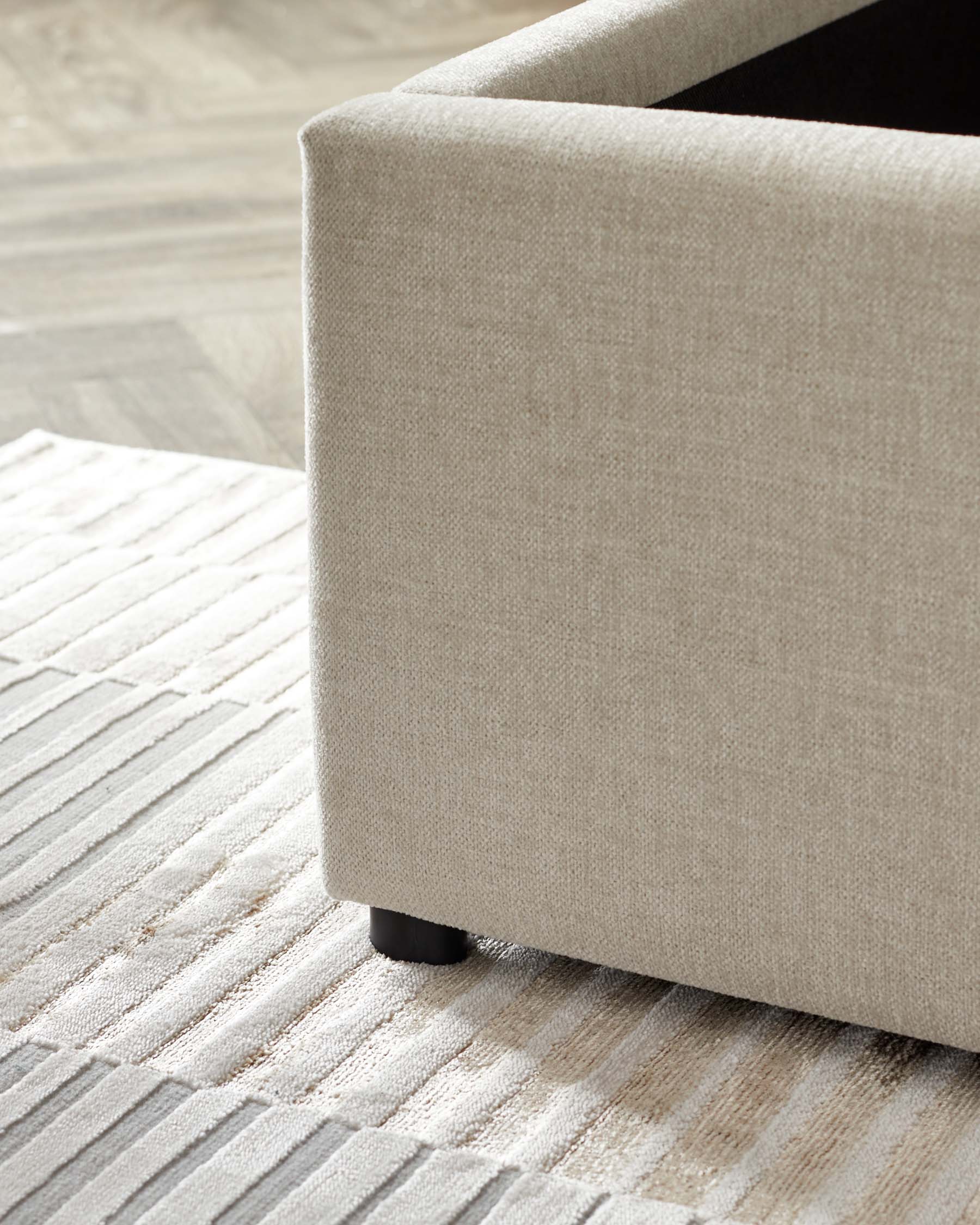 Close-up of a contemporary beige upholstered sofa with a clean-lined silhouette and dark round legs, partially showcased on a textured off-white and beige striped area rug over a wooden floor.