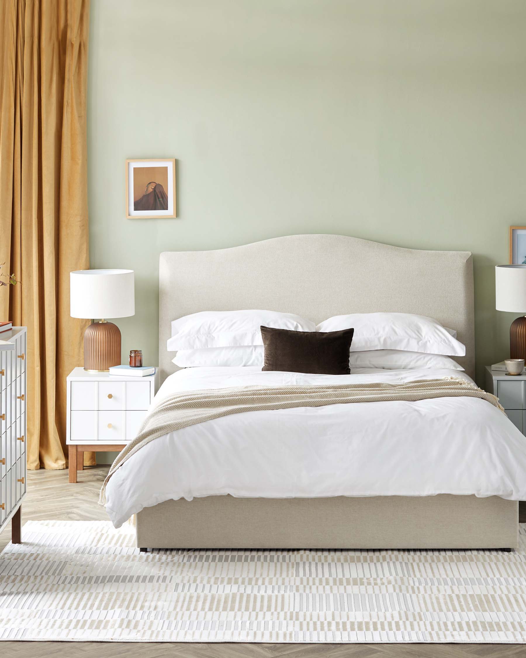 Elegant bedroom furniture setup featuring a neutral-toned upholstered bed frame with a gently curved headboard, flanked by two matching white bedside tables with brass knobs and topped with contemporary white lampshades on ribbed ceramic bases. A textured cream and beige area rug lies beneath the bed, complementing the soothing and sophisticated decor.