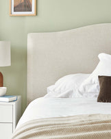 Elegant modern bedroom featuring a large minimalist-style upholstered headboard in a neutral colour, a white nightstand with a drawer, and a classic white table lamp with a cylindrical shade.