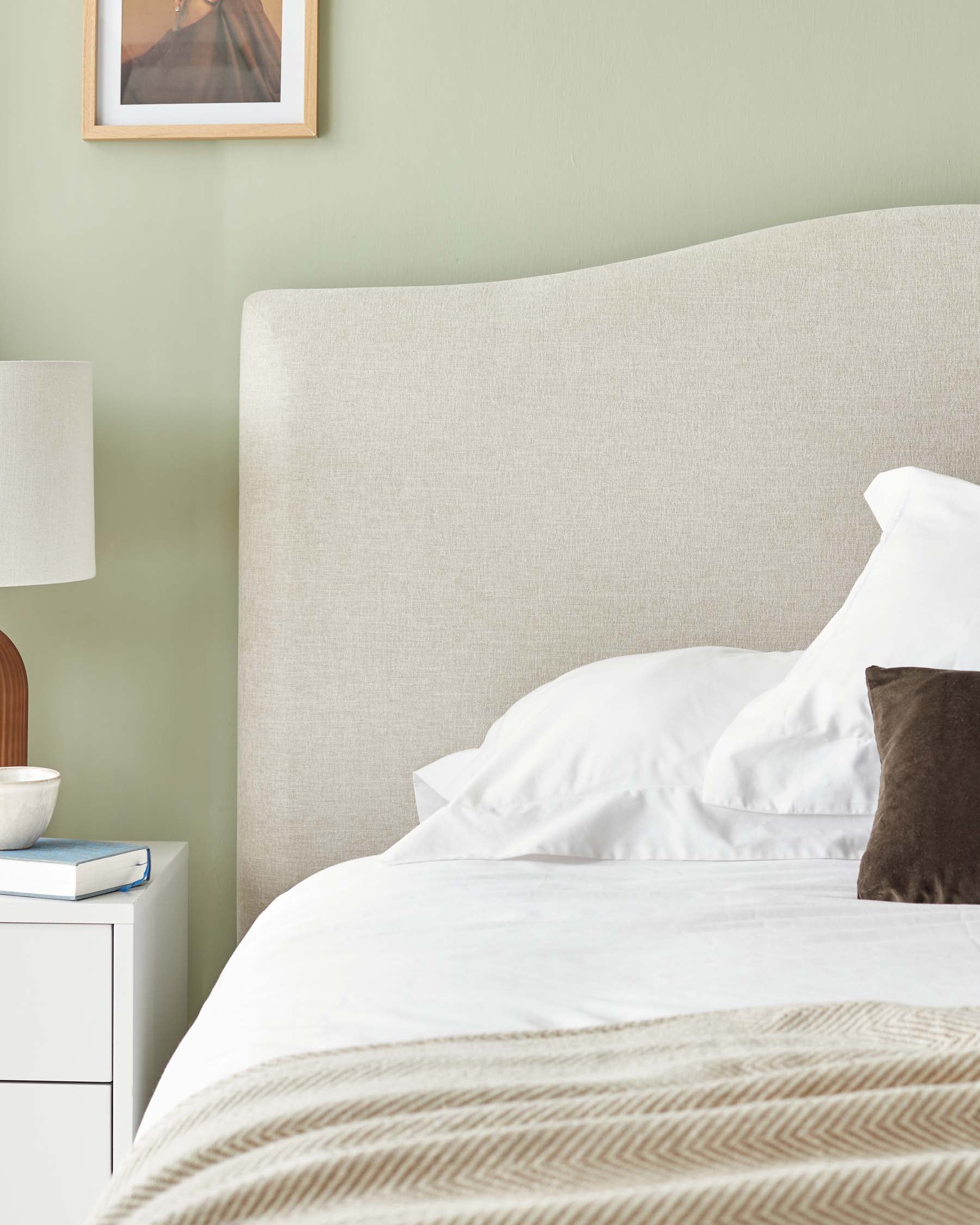 Neutral-toned, curved-top upholstered headboard and a minimalist white bedside table with a single drawer and an open shelf beneath.