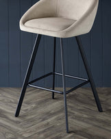 Modern bar stool with a beige upholstered seat and curved backrest on sleek black metal legs with a geometric footrest, showcased against a dark blue panelled wall and wood-patterned flooring.