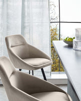 Elegant modern dining chairs with a velvety beige upholstery and sleek black metal legs, set against a backdrop of a contemporary dining table and a large window with sheer curtains, offering a view of autumn foliage.