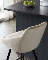 Elegant modern bar stool with a curved, upholstered backrest in a warm beige fabric. The seat is positioned at kitchen counter height and features sleek black metal legs that taper gently towards the floor. The stool is showcased against a dark kitchen island with a marbled countertop, upon which rests a bowl of green apples for a fresh, decorative touch.