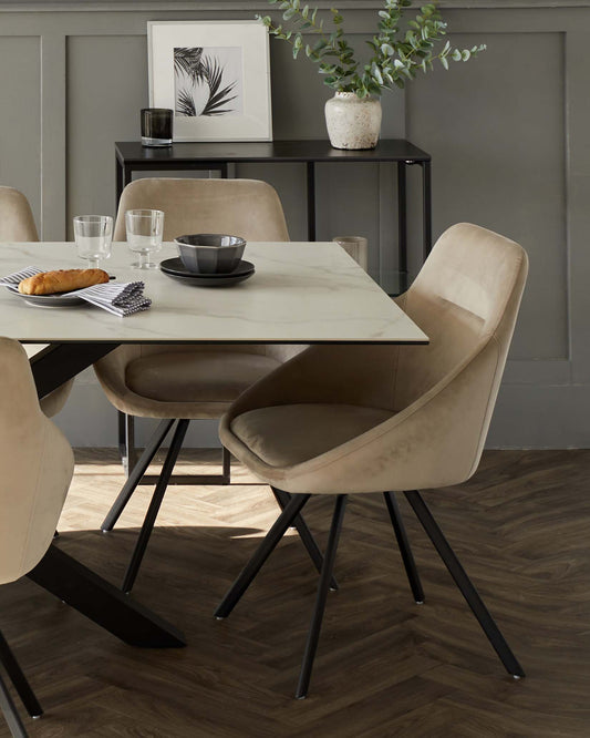 Contemporary dining room set featuring a rectangular marble-top table with a black metal base, complemented by four velvety beige upholstered chairs with black metal legs. A sleek black console table is positioned against the wall in the background.