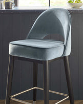 Modern velvet upholstered bar stool with a curved backrest and cushioned seat. Features slim, dark-toned wooden legs with a shiny gold footrest. The chair's fabric is a luxurious deep slate blue, giving it an elegant and sophisticated appearance. Perfect for contemporary kitchen or bar settings.