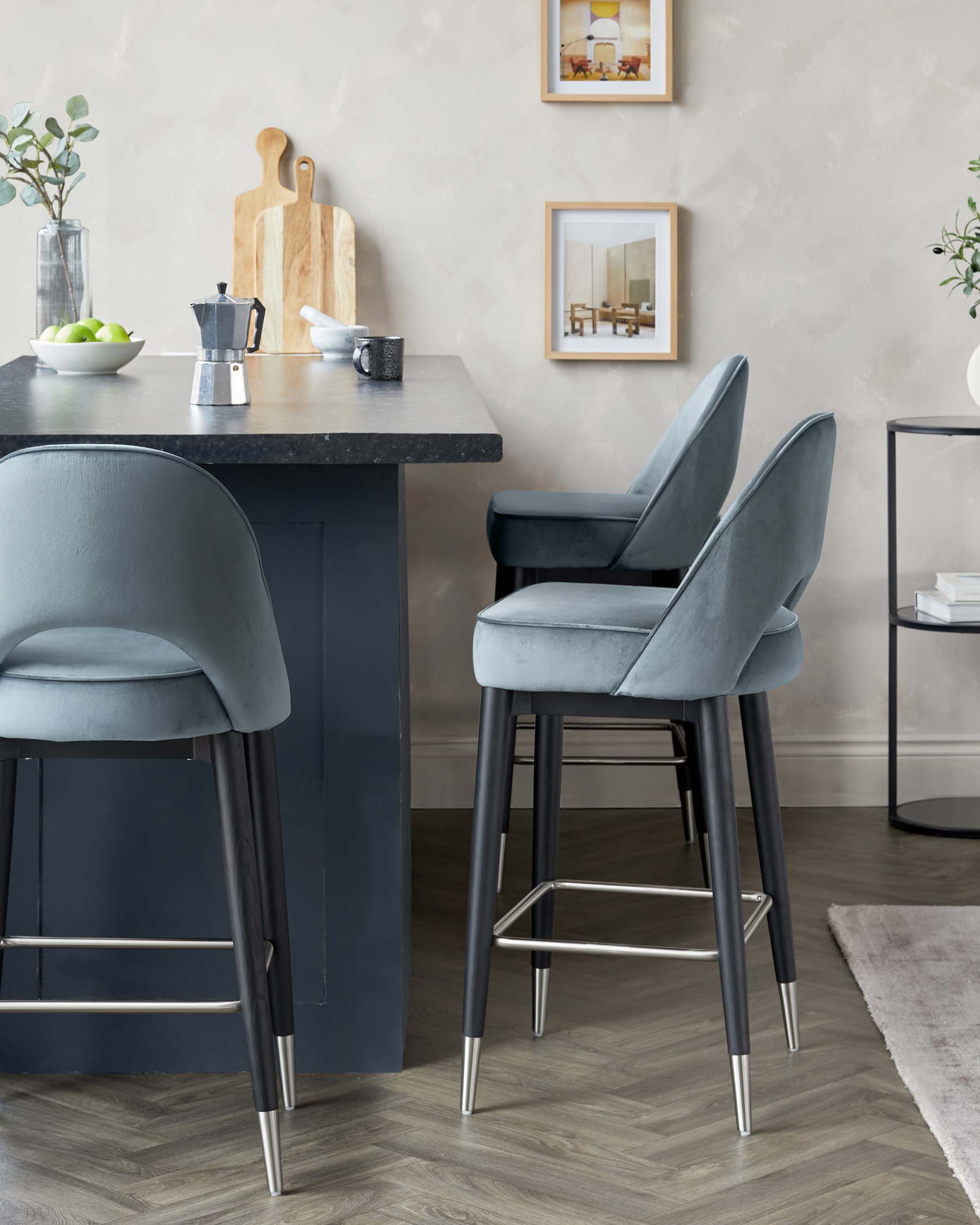 Modern kitchen interior featuring two elegant dark blue upholstered bar stools with sleek metal legs and comfortable back support positioned at a black kitchen island with a light stone countertop. A small side table with a glass surface and metal frame is also visible in the room. The furniture design combines contemporary style with functional comfort, suitable for a modern home aesthetic.