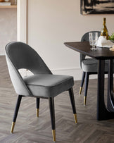 Elegant modern dining furniture featuring a sleek, dark wooden table with a smooth finish and angled legs, paired with a luxurious velvet-upholstered chair in grey with a curved backrest design. The chair boasts black legs that are tipped with stylish gold accents, offering a chic contrast and a touch of sophistication to the aesthetic.