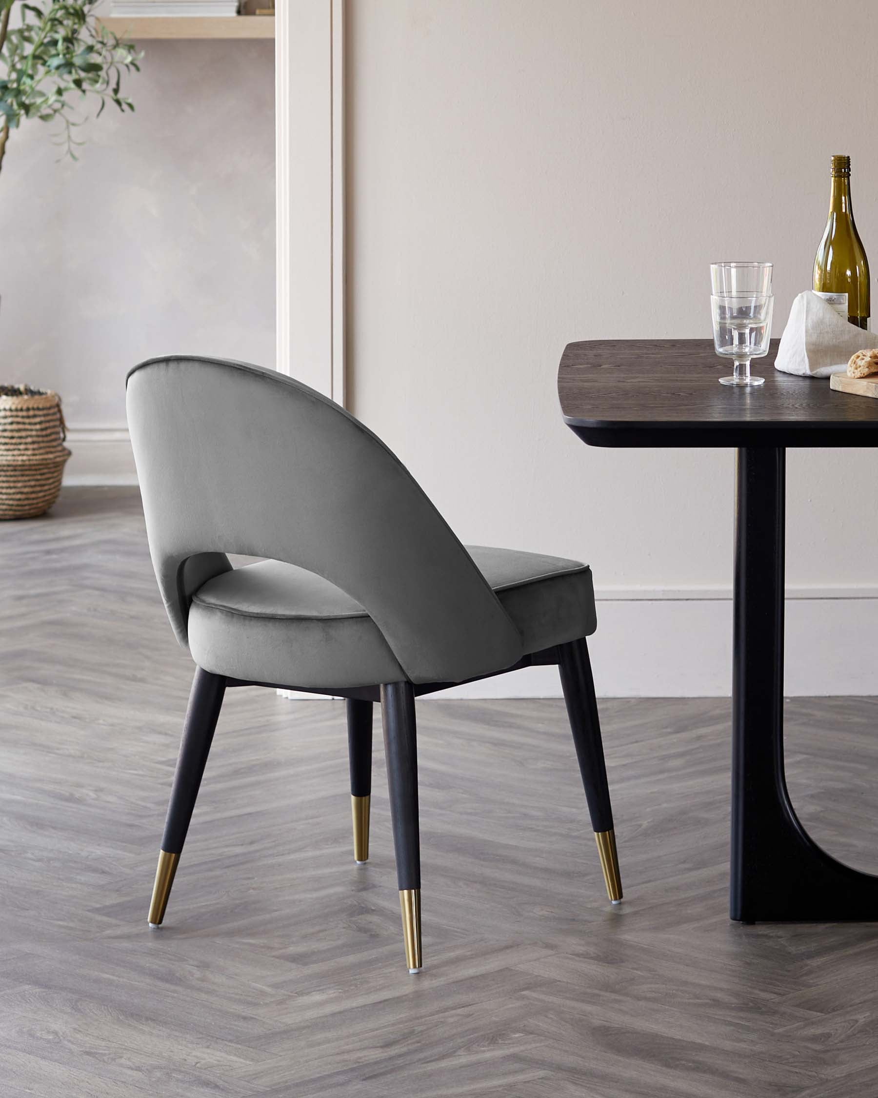 Elegant dining setup featuring a modern velvet upholstered chair with a curved backrest and tapered black legs finished with brass tips, and a minimalistic dark wooden table with a smooth, round-edged rectangular top and solid cylindrical legs.