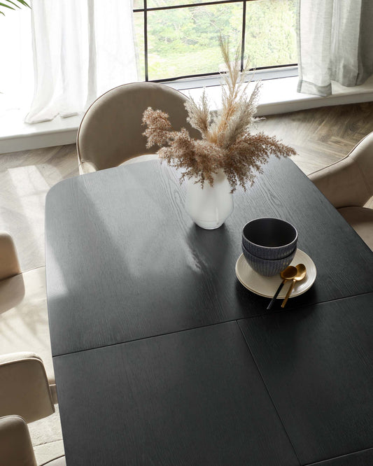 Stylish black table with smooth finish, cozy beige chairs, a white vase with dried foliage, and contemporary dishware.