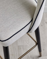 Cream upholstered chair with black trim, featuring a sleek black wooden leg and gold metal accents for a modern touch.