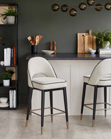 Two modern bar stools with cream upholstery and black legs, accented with gold tips, beside a stylish kitchen counter.