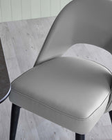 Modern, minimalist-style chair featuring a sleek, curved design with a padded grey leatherette seat and backrest, mounted on slender black metal legs. The chair offers a sophisticated aesthetic with a comfortable, supportive seating experience.