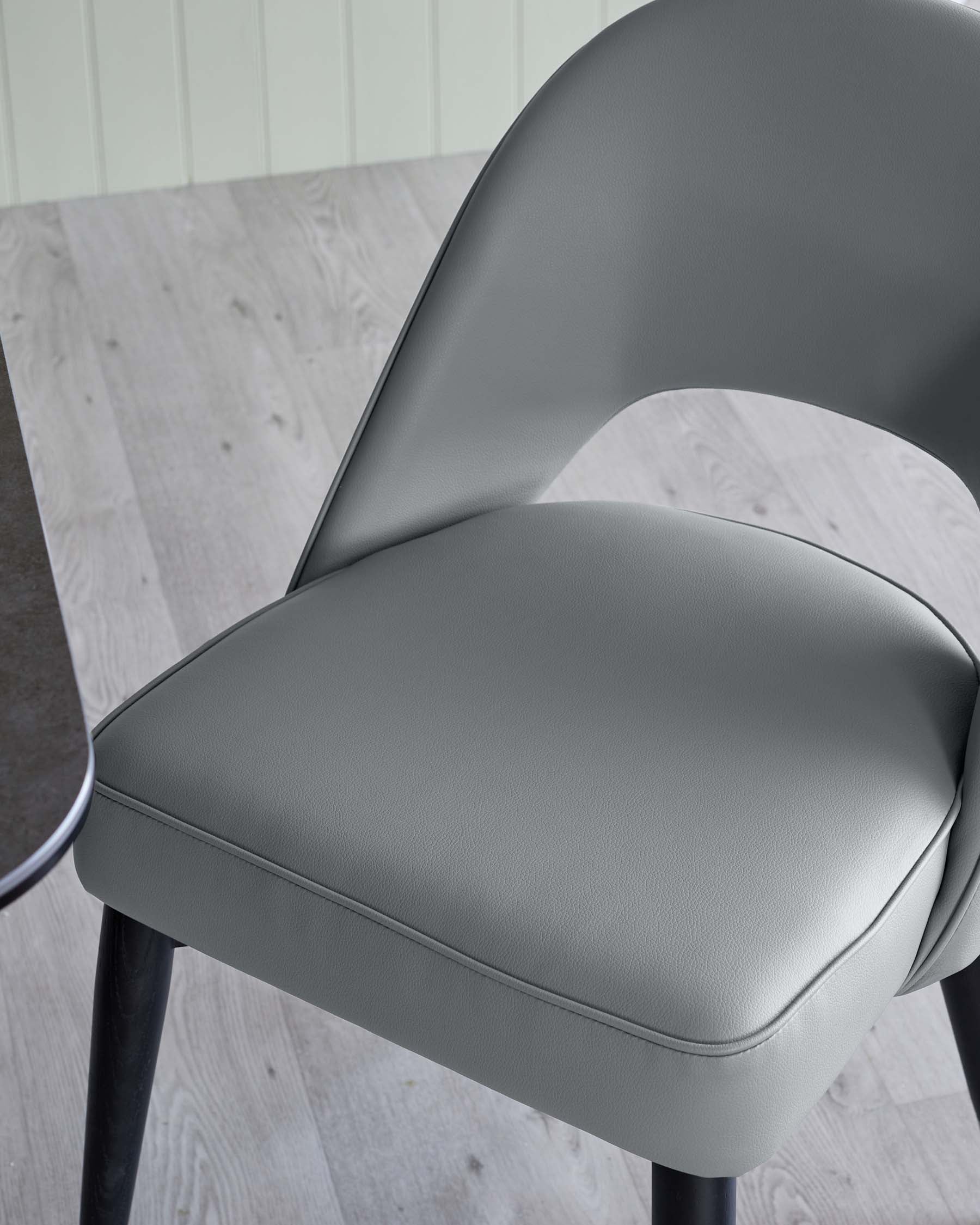 Close-up of a modern chair with a stylish, curved backrest and comfortable, padded seat upholstered in smooth grey faux leather, supported by sleek black metal legs. The flooring features light wood laminate, with a soft pastel wall in the background providing a calm, contemporary setting.