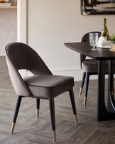 Elegant modern dining chair with dark grey upholstery and black legs featuring gold-coloured tips, alongside a contemporary black dining table with a bottle of wine and a glass set upon it.