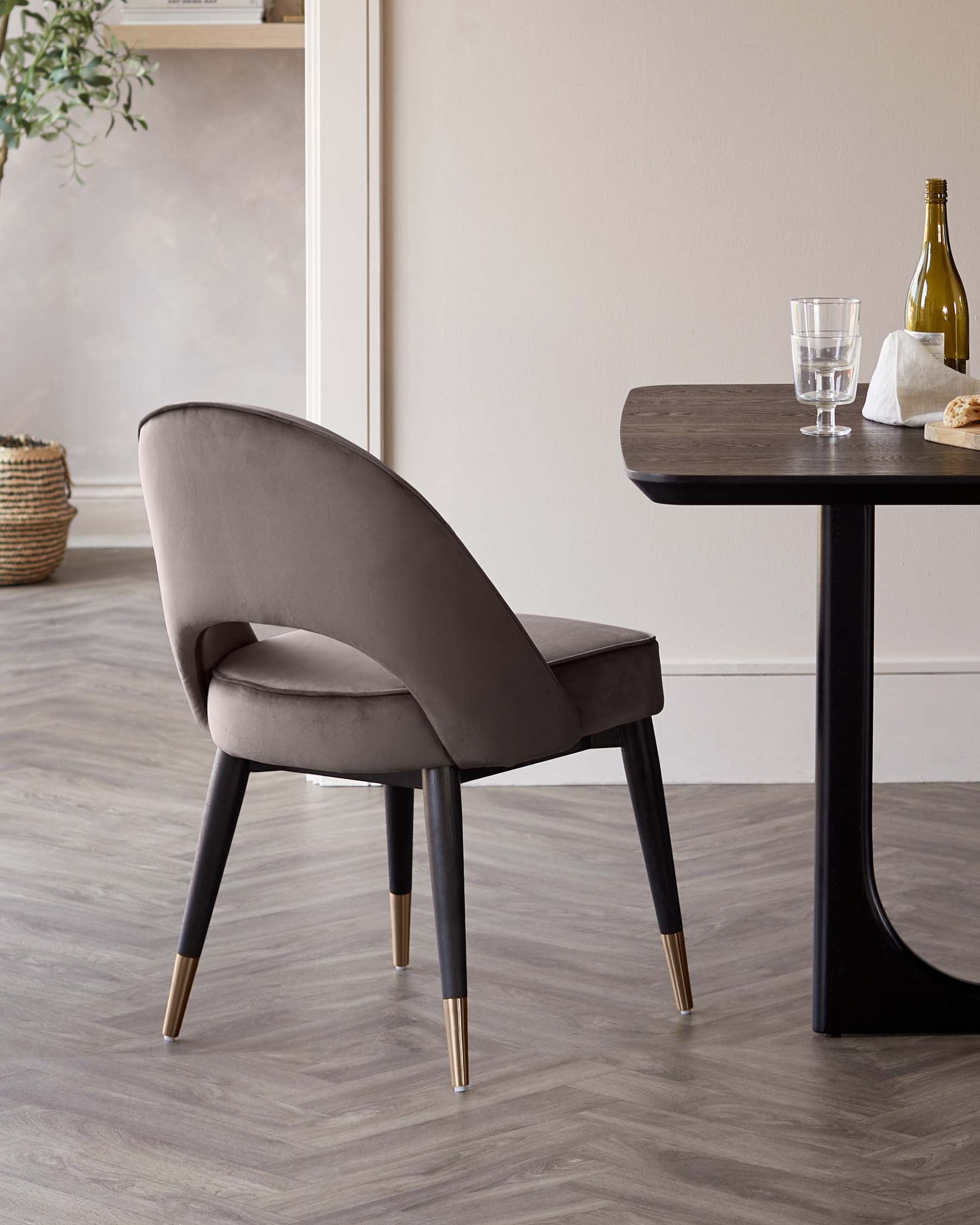 A contemporary dining chair with a curved backrest and grey upholstery sits next to a sleek dark-toned wooden dining table. The chair features black legs tipped with brass caps, complementing the table’s minimalist silhouette.