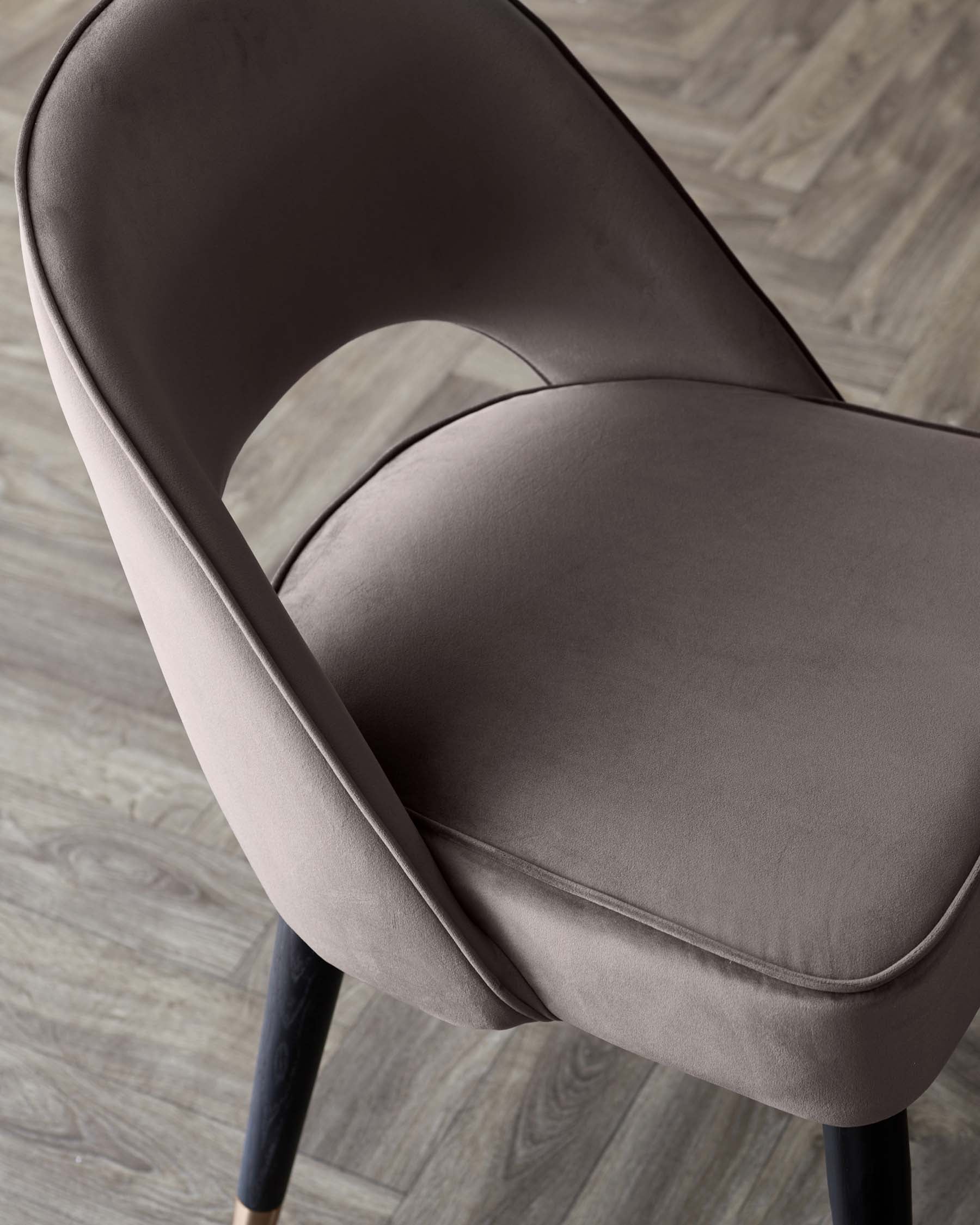 Elegant modern chair with a smooth taupe upholstery and dark wooden legs, featuring a curved backrest and a plush seat, set on a herringbone pattern wooden floor.