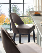 Elegant modern dining chair with a smooth, grey upholstered seat and a curved backrest, supported by slender, dark wood legs, showcasing a minimalist design. Positioned against a backdrop of floor-to-ceiling windows with a view of autumn foliage.