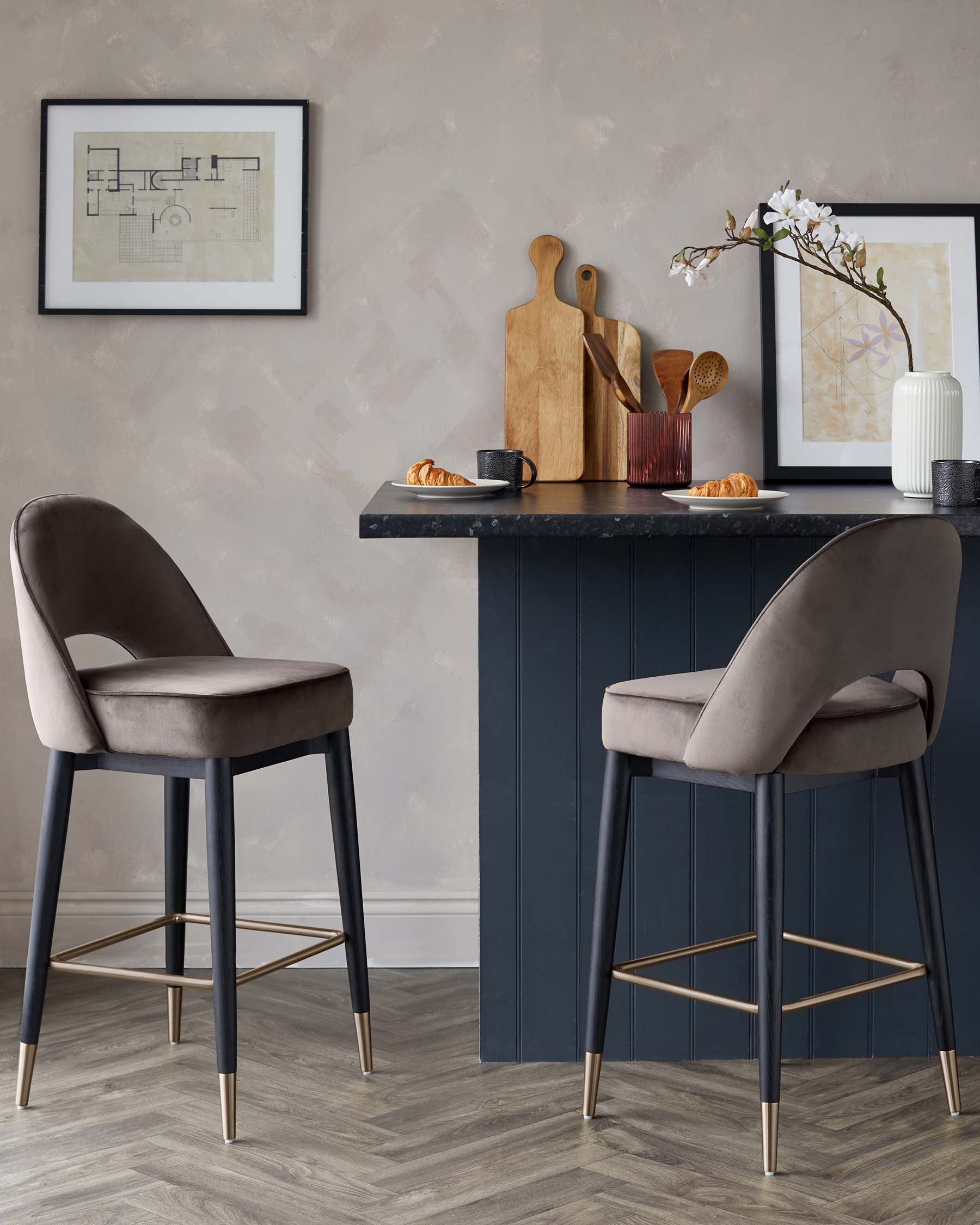 Two elegant contemporary barstools with plush, velvet-upholstered seats in a rich taupe colour, featuring curved backrests and slim, tapered legs in a dark finish with metallic gold footrests and accents. A sophisticated blue-painted bar counter is also visible, showcasing a sleek, minimalist design.