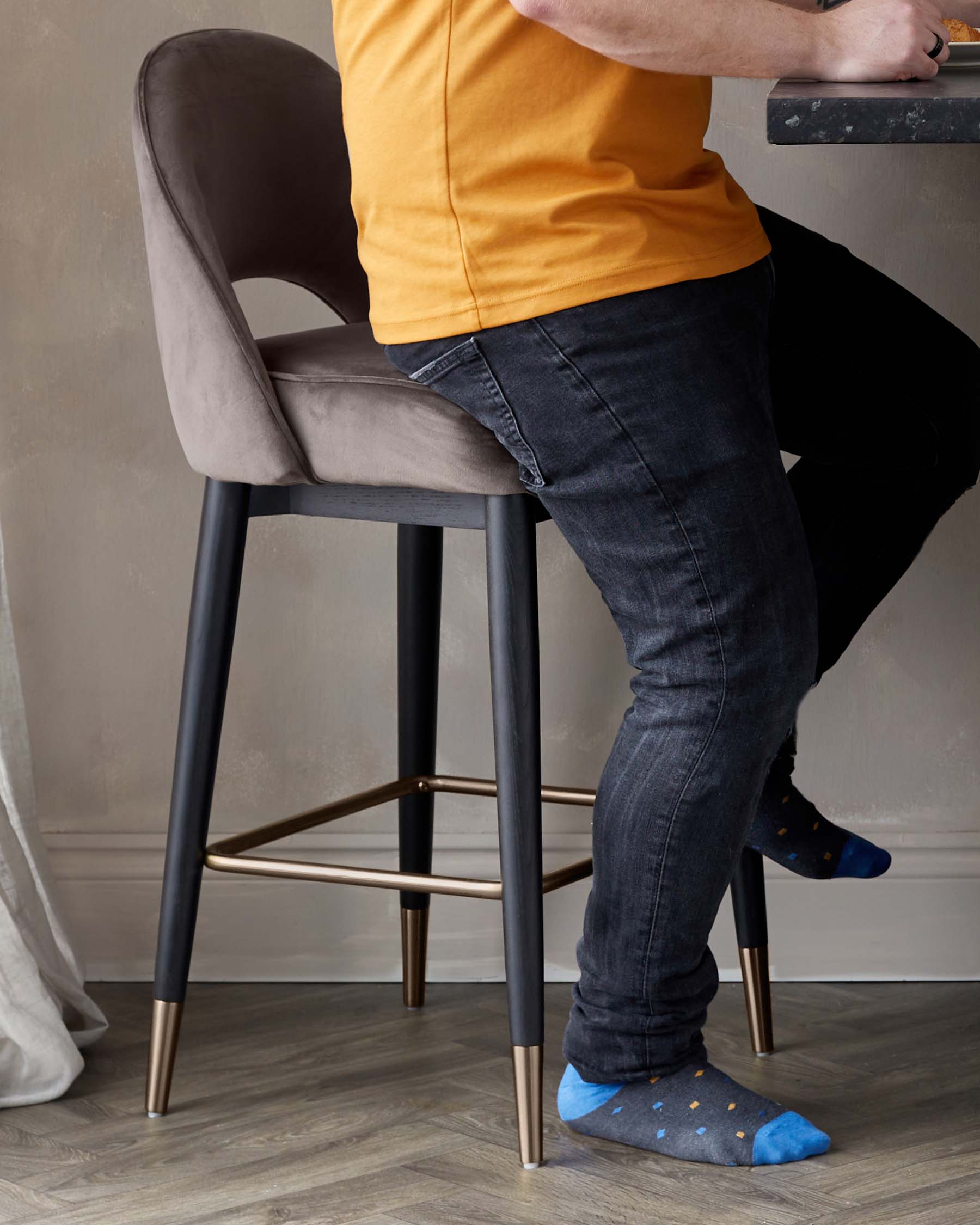 A modern bar stool with a taupe-coloured upholstered seat and backrest, supported by four slender black metal legs with elegant gold-coloured tips and a matching gold footrest.