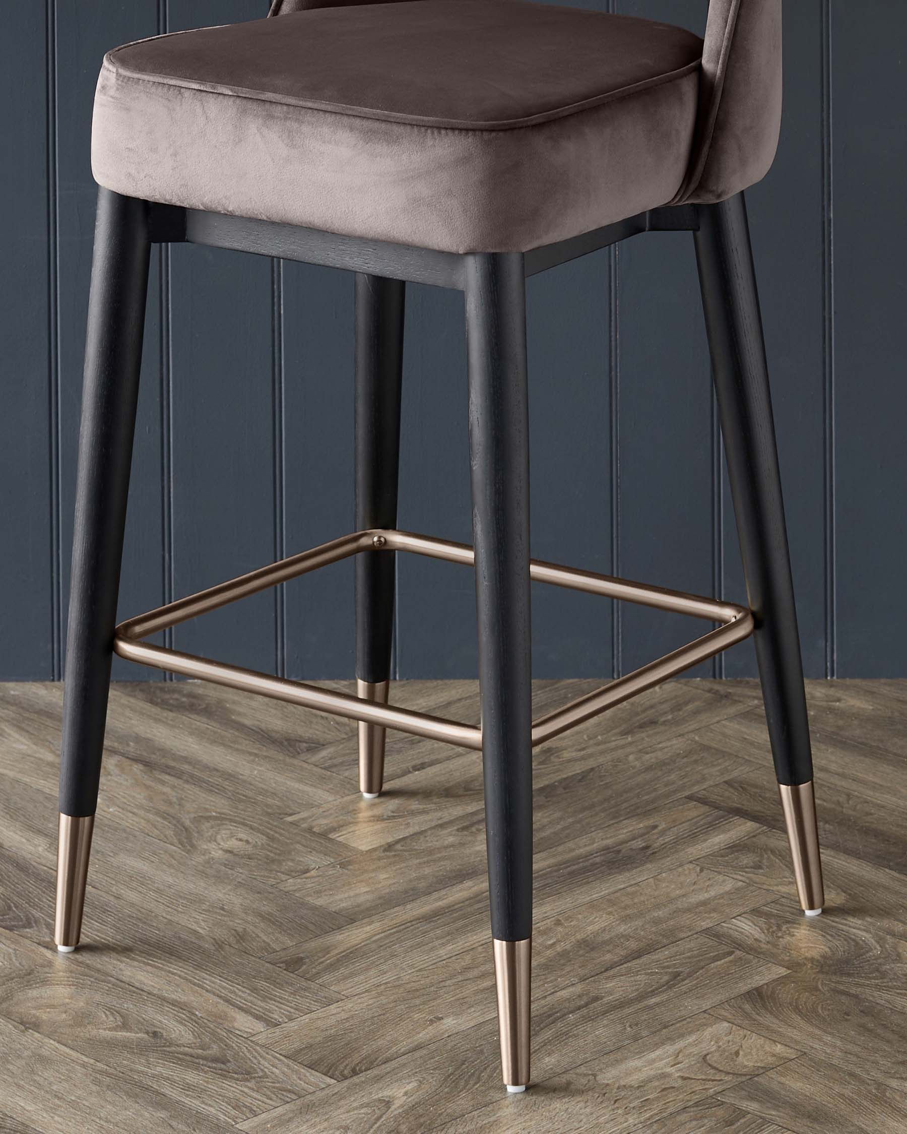 Elegant modern bar stool with a smooth, grey velvet seat cushion and backrest on a sleek, black frame with contrasting metallic gold footrest and leg tips, set against a dark wainscot wall on a herringbone wooden floor.