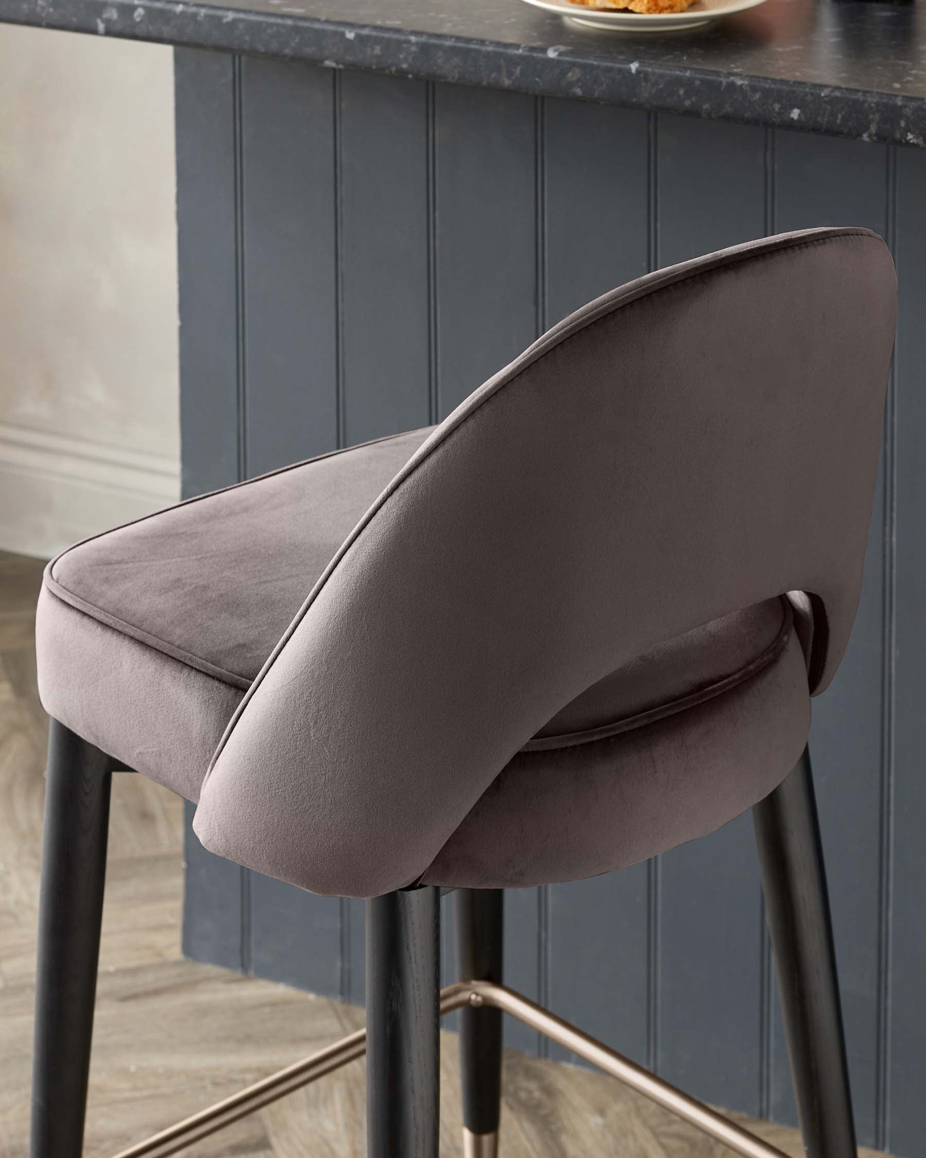 Elegant modern bar stool with a curved backrest and plush, velvety upholstery in a muted taupe shade. Features sleek black metal legs accented with gold footrests, juxtaposed against a dark panelled wall and herringbone wood floor.