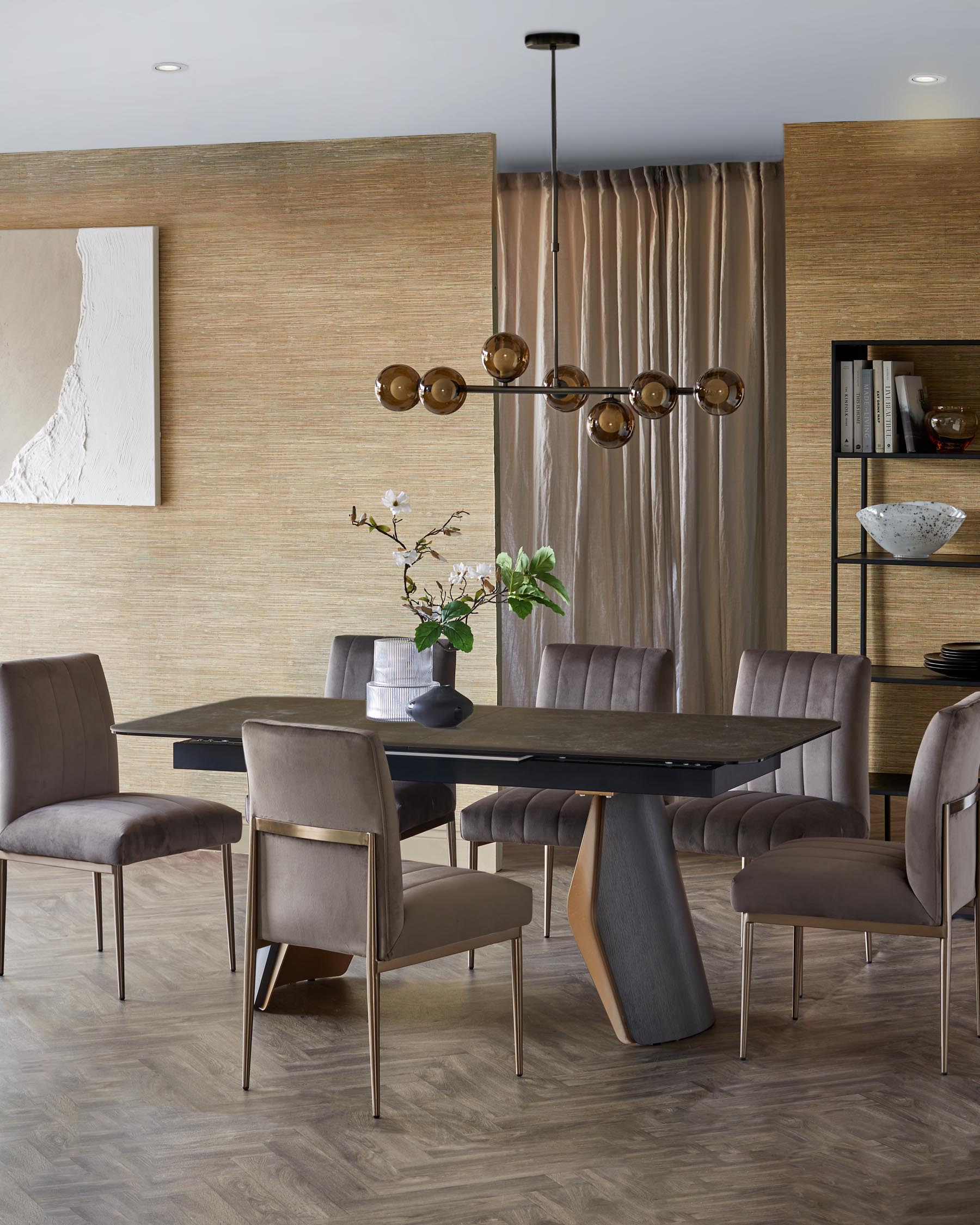 A modern dining table with a unique base, accompanied by upholstered chairs with gold accents, a stylish chandelier, and decor.