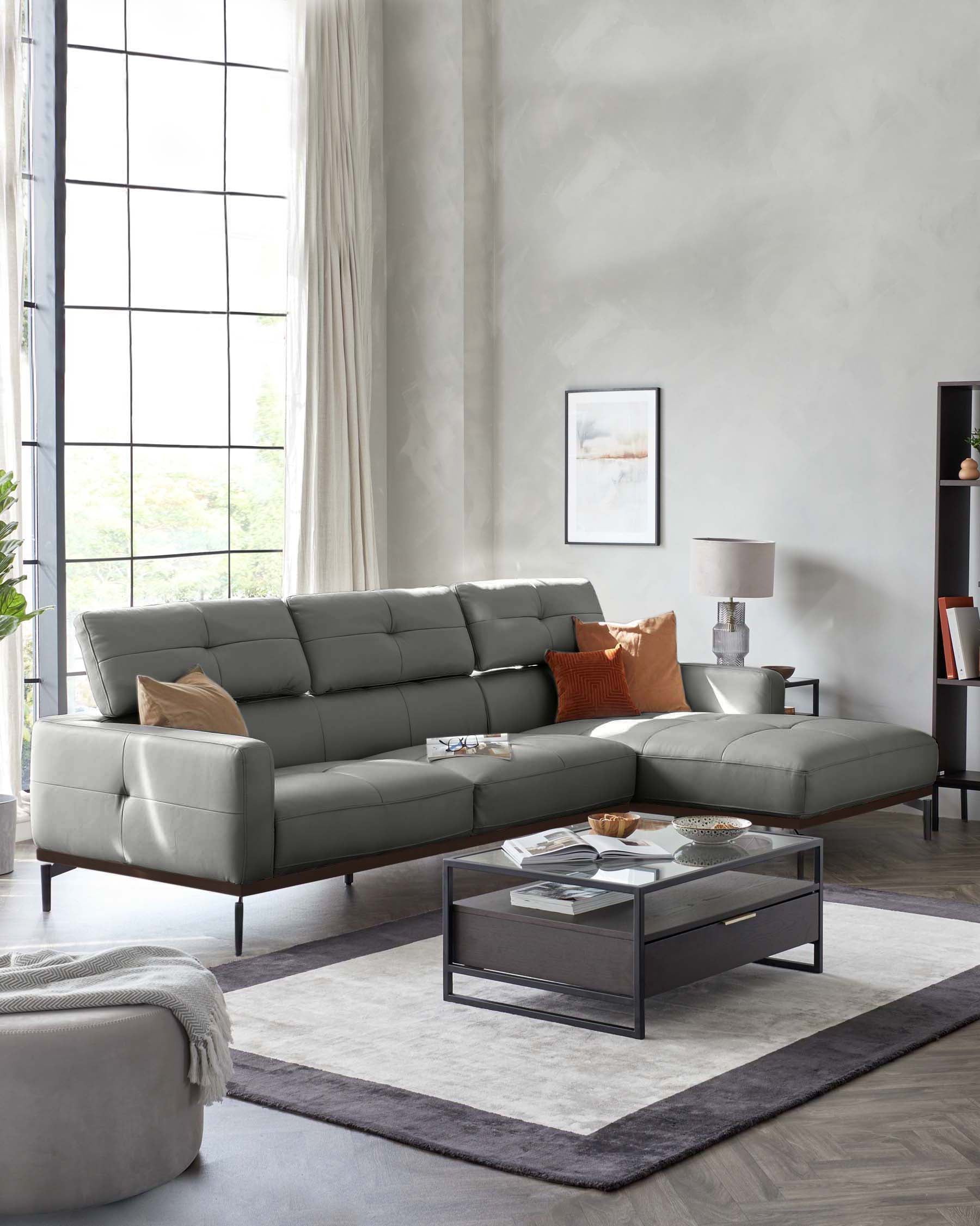 Modern living room showcasing a grey L-shaped sectional sofa with plush cushions and a chaise on one end. In front of it is a sleek rectangular glass-top coffee table with a metal frame and a lower shelf. A soft beige ottoman sits off to the side, complementing a neutral-toned area rug underneath the furniture.