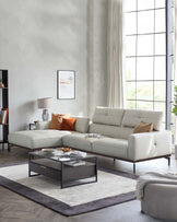 Elegant light grey L-shaped sectional sofa with slim metal legs, paired with a modern square black coffee table with a glass top and matching black frame. A plush grey and white area rug underlines the set, and a round grey ottoman sits in the foreground.