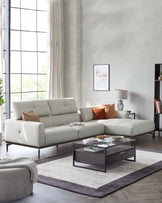 A modern living room featuring a sleek, light-coloured leather L-shaped sectional sofa with clean lines and slender metallic legs. In front of it rests a contemporary rectangular glass-top coffee table with a dark wooden shelf and a minimalist metal frame. Adjacent to the sofa is a round, matching upholstered ottoman. The ensemble is arranged on a large two-tone area rug with a plush texture, coordinating harmoniously with the room's neutral colour palette.