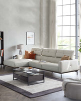 Elegant modern living room setup with a light grey L-shaped sectional sofa featuring plush back cushions and a chaise lounge on the right side. A sleek, square glass-top coffee table with a black frame and lower shelf sits atop a two-tone grey area rug. A muted grey cylindrical ottoman is positioned to the side. The room is accented with a dark wood bookcase and various decor elements, creating a chic and sophisticated interior space.