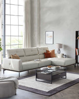 Modern living room featuring a light beige L-shaped sectional sofa with plush cushions, accompanied by a round, fabric-upholstered ottoman in a matching shade. At the centre lies a contemporary, rectangular coffee table with a black metal frame and a glass top, alongside a grey and white area rug beneath.