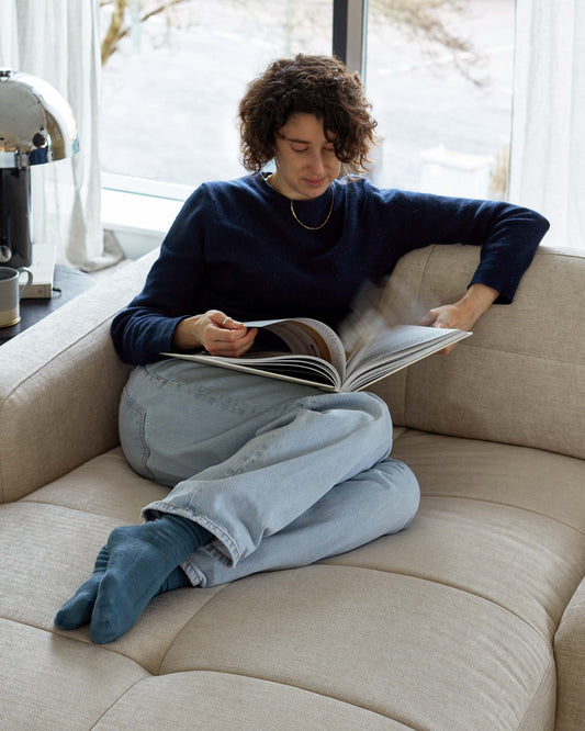 A beige fabric sofa with a modern and minimalist design. The sofa has a clean, square silhouette with plush, wide cushions for seating and a padded, structured backrest. It stands in a well-lit space by a large window, inviting relaxation and comfort.