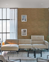 Modern minimalist living room interior with a neutral-toned sectional sofa featuring clean lines and a plush look. In front of the sofa is a round glass coffee table with a black metal base. The room also has a matching armchair with a simple metal frame, located at the edge of the frame. The furniture is arranged on a light textured area rug, complementing the contemporary aesthetic of the space.