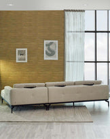 A modern beige sectional sofa with a chaise on one end, featuring a sleek design with clean lines, tufted seat cushions, and six cylindrical black pillows. It rests on a thin dark metal base that provides a floating appearance. The sofa is placed on a textured light grey area rug, against a golden textured wall in a contemporary room with large windows draped with sheer white curtains.