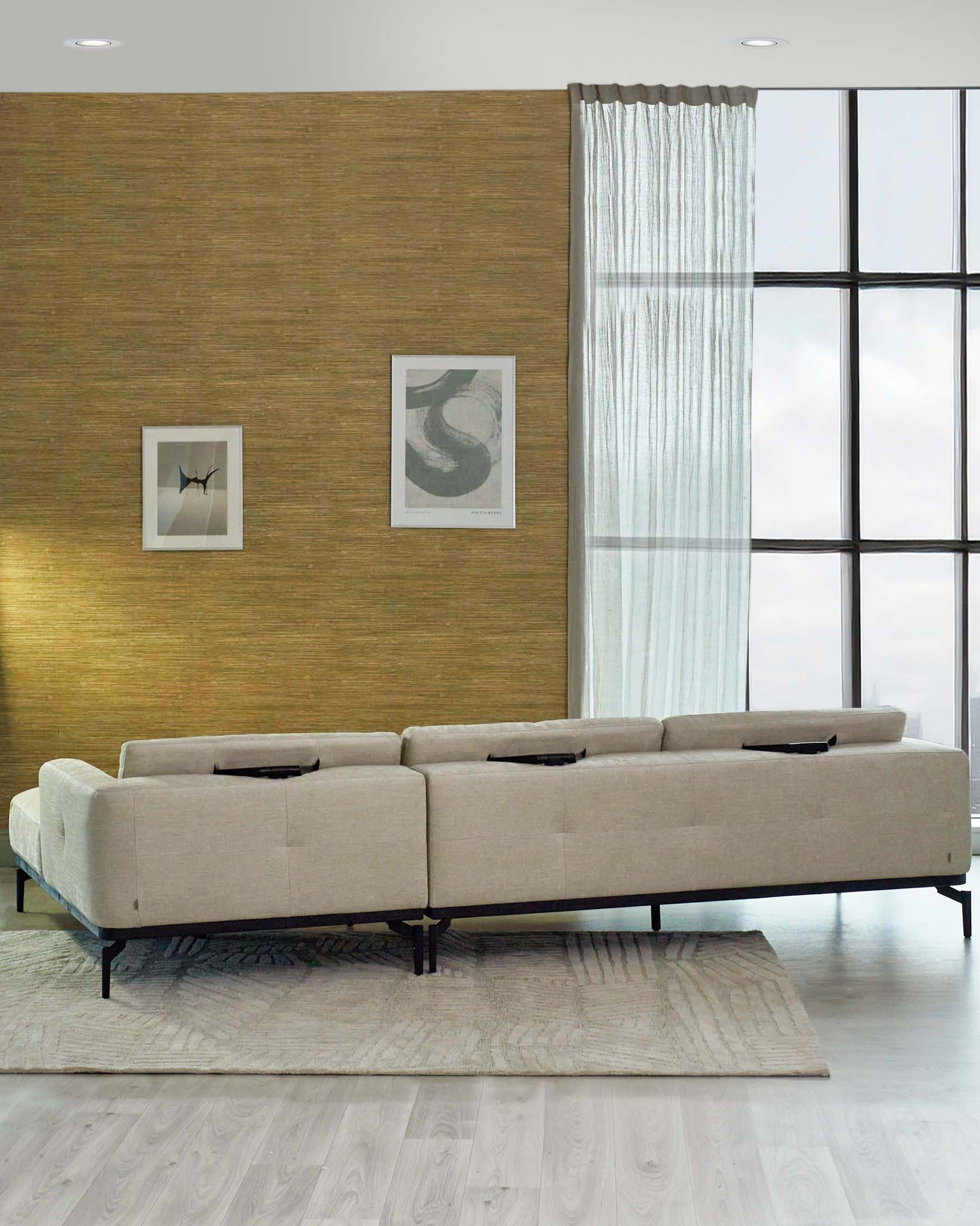 A modern beige sectional sofa with a chaise on one end, featuring a sleek design with clean lines, tufted seat cushions, and six cylindrical black pillows. It rests on a thin dark metal base that provides a floating appearance. The sofa is placed on a textured light grey area rug, against a golden textured wall in a contemporary room with large windows draped with sheer white curtains.