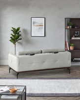 Modern light grey upholstered sofa with tufted back cushions and dark tapered wooden legs, paired with a sleek low-profile black coffee table featuring an open shelf, set on a layered grey area rug. A dark wooden bookshelf with various decorative items and books completes the contemporary look.