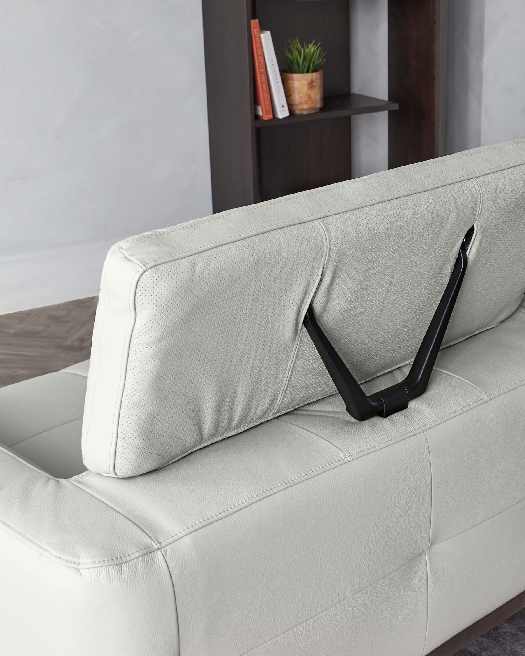 A modern, off-white leather sofa with a structured and plush backrest and seat. There’s a sleek black metal frame that curves around the back adding a contemporary touch. The sofa is set against a room with a dark multishelved bookcase filled with decor items, suggesting a chic and minimalist aesthetic.