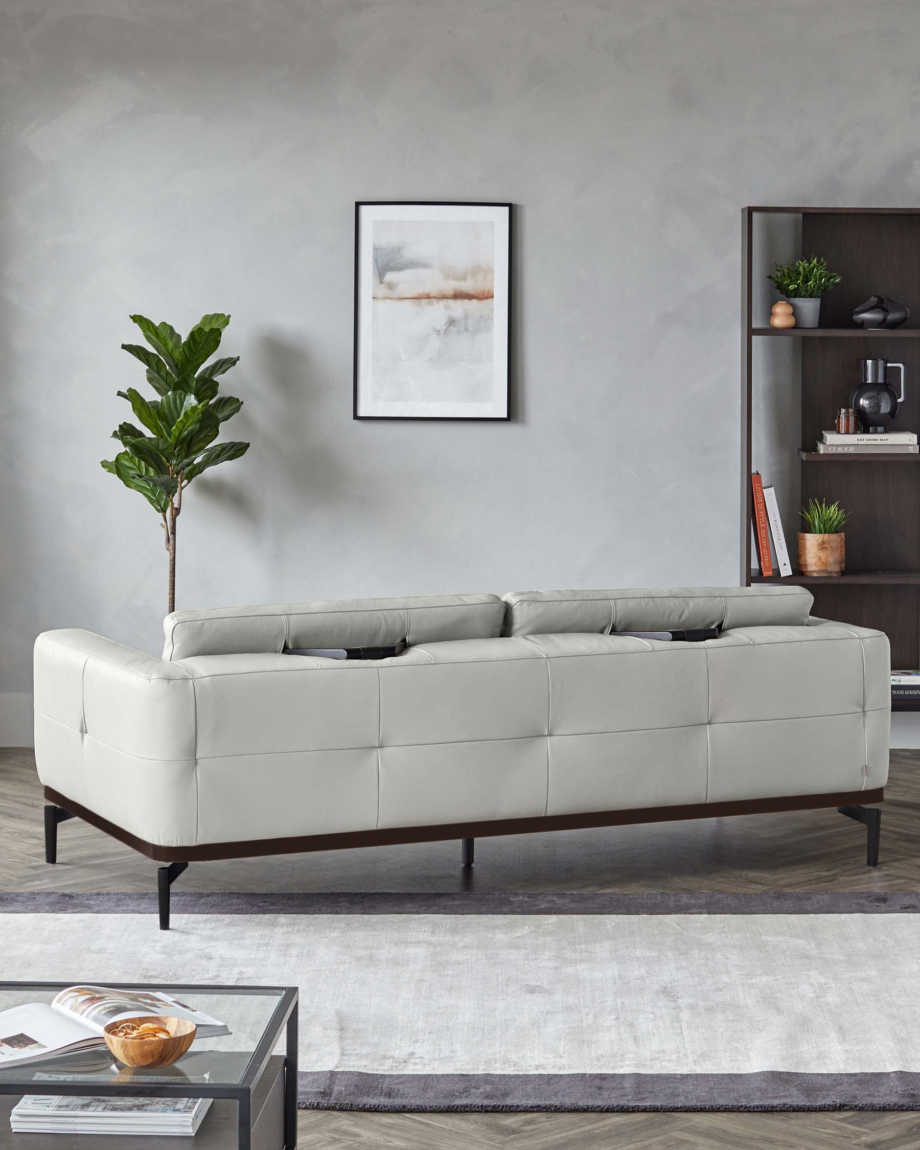 Modern light grey upholstered sofa with tufted back and seat cushions, elevated on slim, dark wooden legs. To the right, there is a dark wood bookshelf with various decorative items and books, and at the front, a low-profile grey coffee table with a glass top over magazines. A white and grey area rug is underneath, and a potted fiddle leaf fig tree stands to the left.