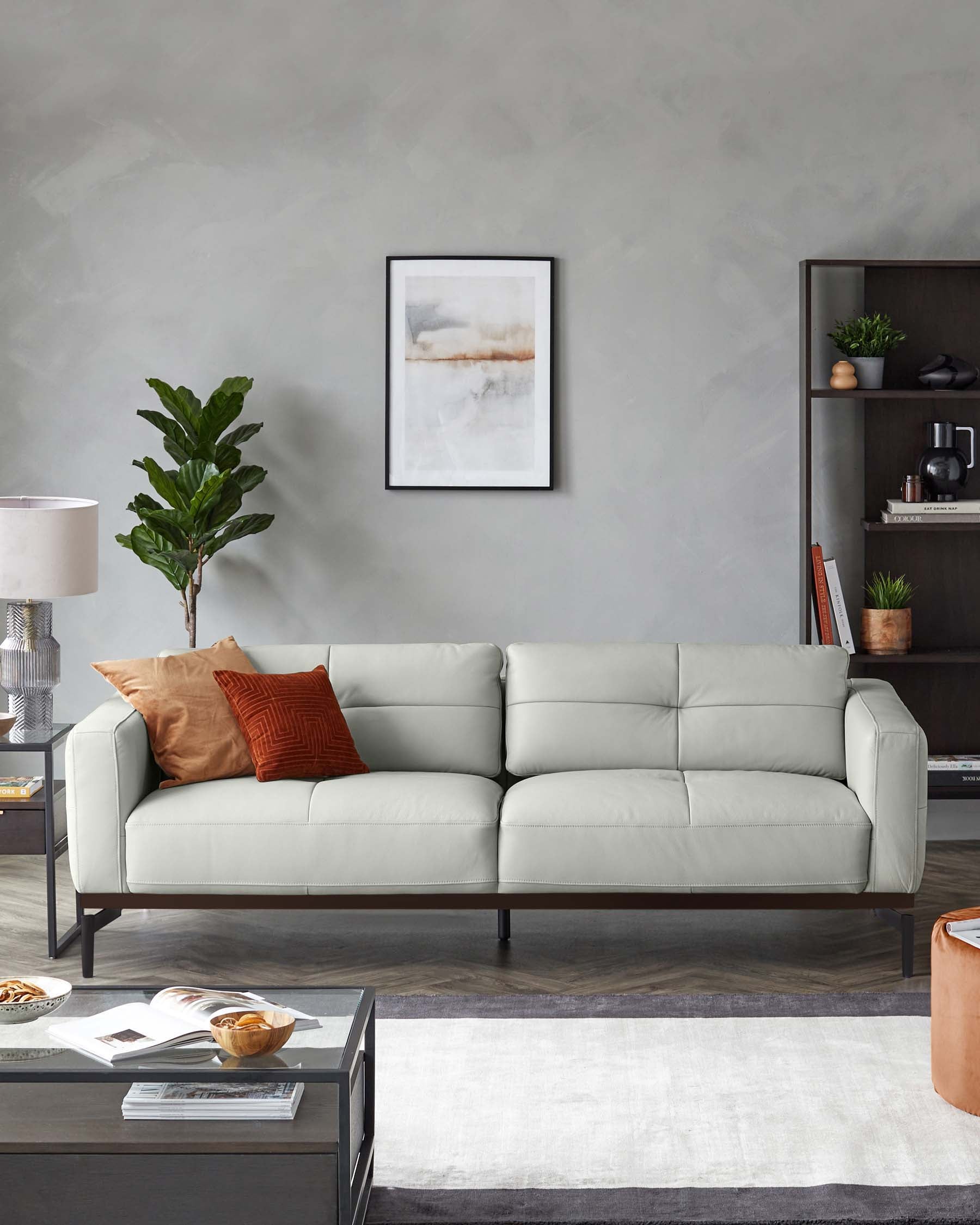 A contemporary light grey three-seater leather sofa with clean lines and dark tapered legs, accompanied by a modern dark wood side table and a coordinating coffee table. Accessories include a white and grey area rug, decorative pillows, and a potted indoor plant adding a touch of greenery to the space. A framed abstract artwork hangs on the wall above the sofa, and a shelf with books and small plants contributes to the modern aesthetic.