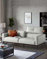 Elegant modern living room featuring a light grey upholstered sectional sofa with a sleek silhouette and thin metal legs, complemented by an array of textured cushions. In front, a contemporary glass-top coffee table with a matte black frame sits atop a monochromatic area rug with geometric patterns. A slim floor lamp with a white shade and a dark wood bookshelf adorned with decorative objects and potted plants add warmth to the scene.