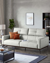 A modern light gray sofa with tufted cushions, accented by an orange pillow, paired with a glass coffee table and decorative plant.