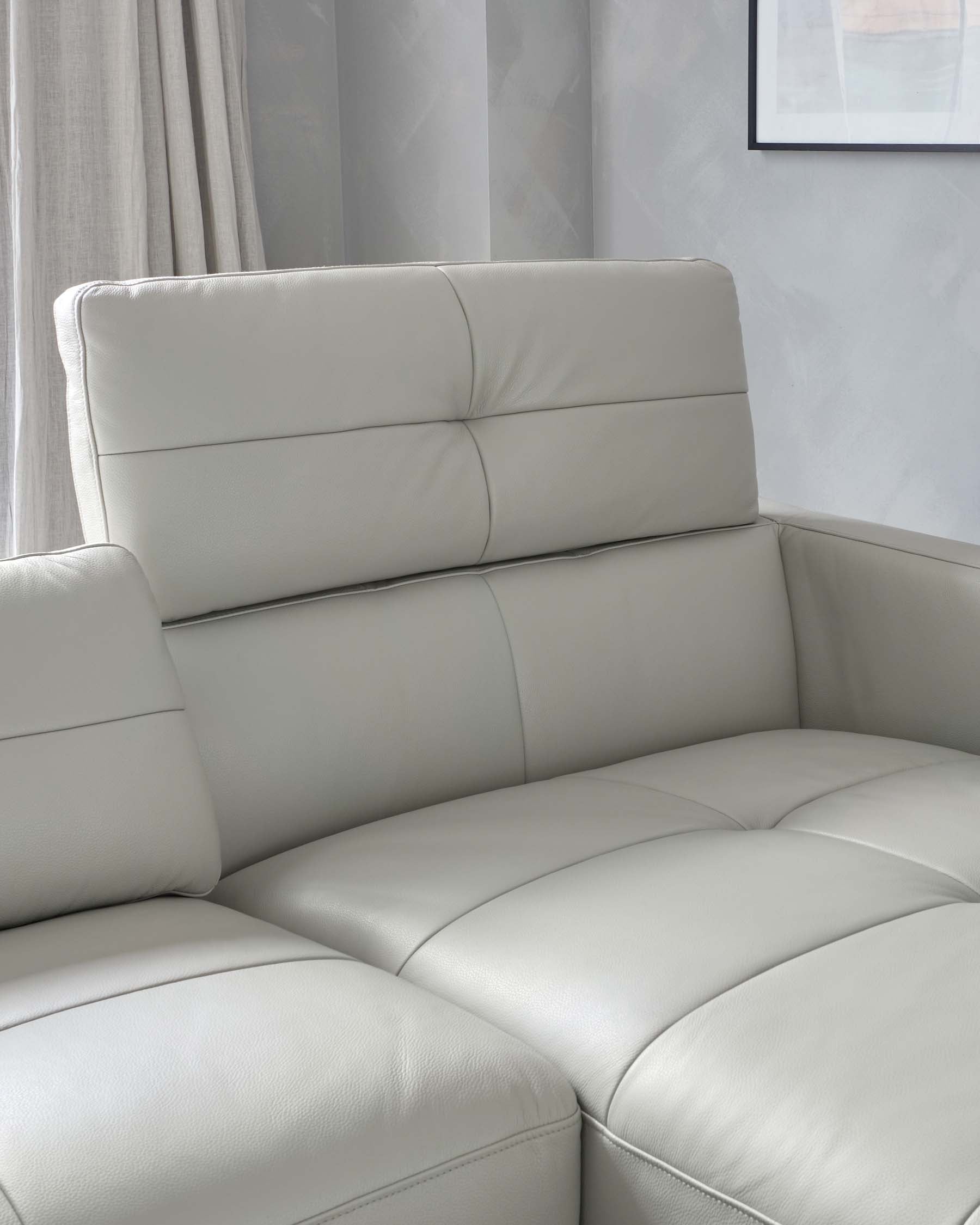 Modern light grey leather sectional sofa with clean lines and a plush backrest, featuring deep padded seats, situated in a well-lit room with soft grey curtains in the background.