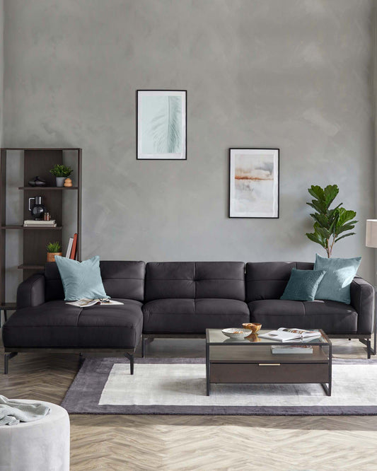 Modern black sectional sofa with teal pillows, glass-top coffee table with drawer, and stylish bookshelf against a gray wall.