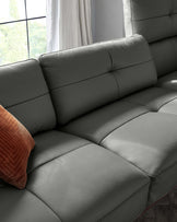 Modern grey leather sofa with plush cushions and a single terracotta-coloured throw pillow. The sofa is shown in a room with large windows draped with sheer curtains, overlooking greenery outside.