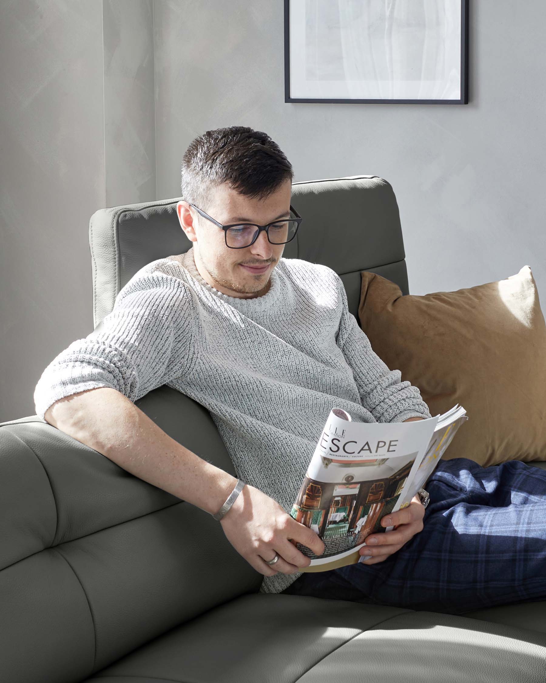 A contemporary grey leather armchair with a high back and a cushioned seat, complemented by a soft brown throw pillow on one side. The armchair is positioned in a naturally lit corner with a simple, framed artwork hanging on the wall above.