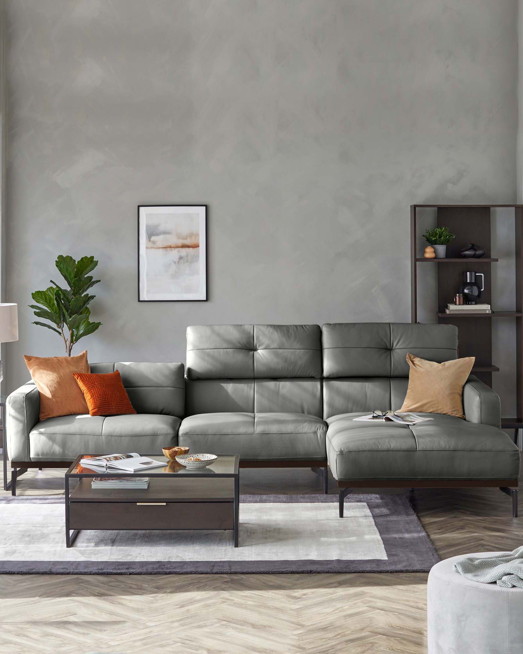 A modern living room featuring a sleek grey L-shaped sectional sofa with clean lines and plush cushions, complemented by orange decorative pillows. In front of the sofa is a contemporary rectangular coffee table with a glass top and dark wood base, displaying books and decorative items. A dark shelving unit stands to the right, subtly matching the coffee table and showcasing minimalistic decor and houseplants. The room is completed with a soft-toned area rug underneath.