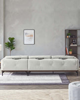 Modern light grey upholstered sectional sofa with clean lines and simple metal legs, set on a white and grey area rug, complemented by a minimalist dark wood shelving unit displaying decorative items. A potted plant and framed abstract wall art add a touch of greenery and colour to the contemporary setting.